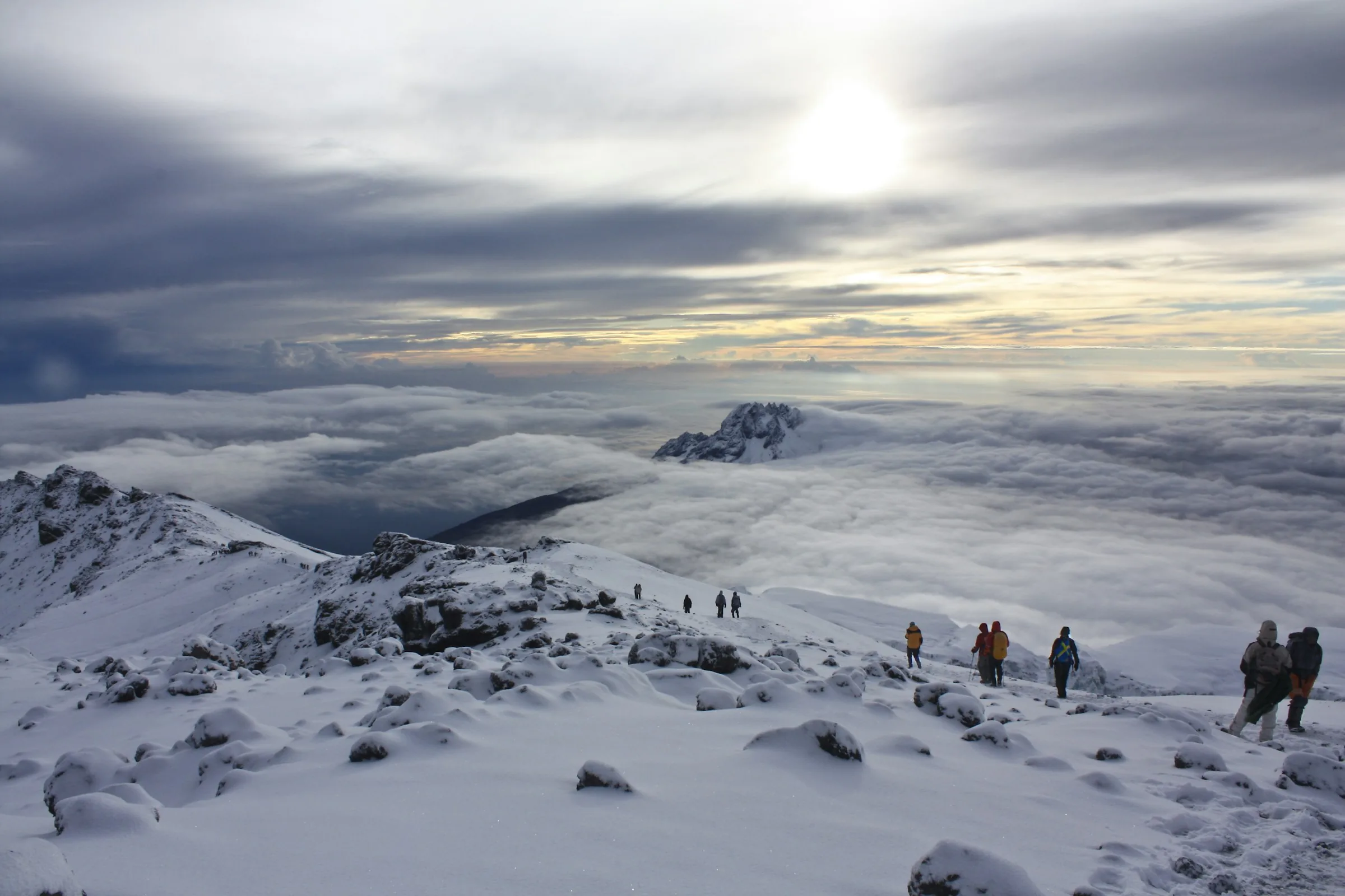Dawn above the clouds high on Kilimanjaro, climbers crossing fresh snow near Stella Point with Mawenzi rising through the cloud